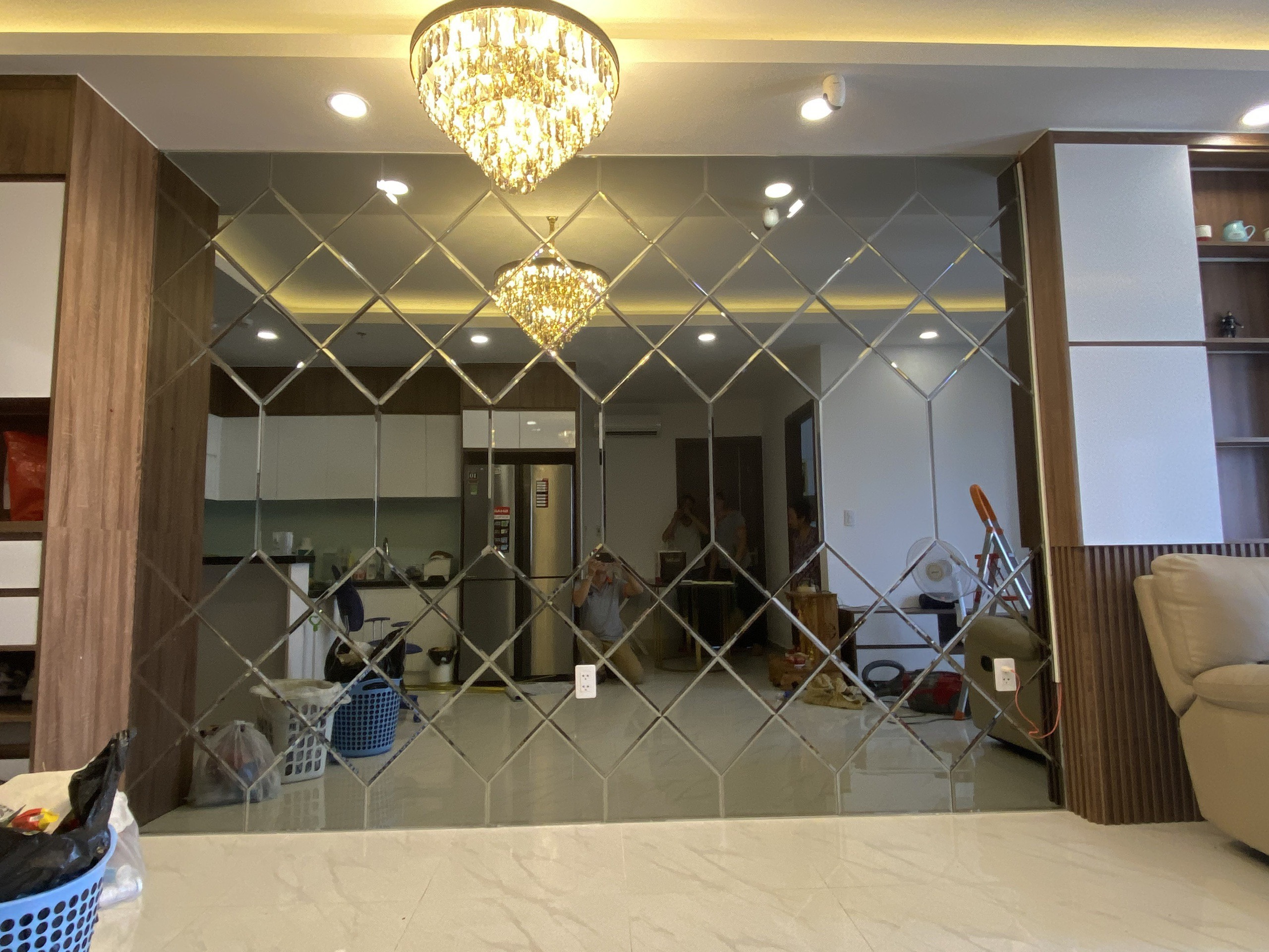 Mảng gương ghép quả trám mài vát phủ kín tường phòng khách căn hộ, phản chiếu đèn chùm và khu bếp