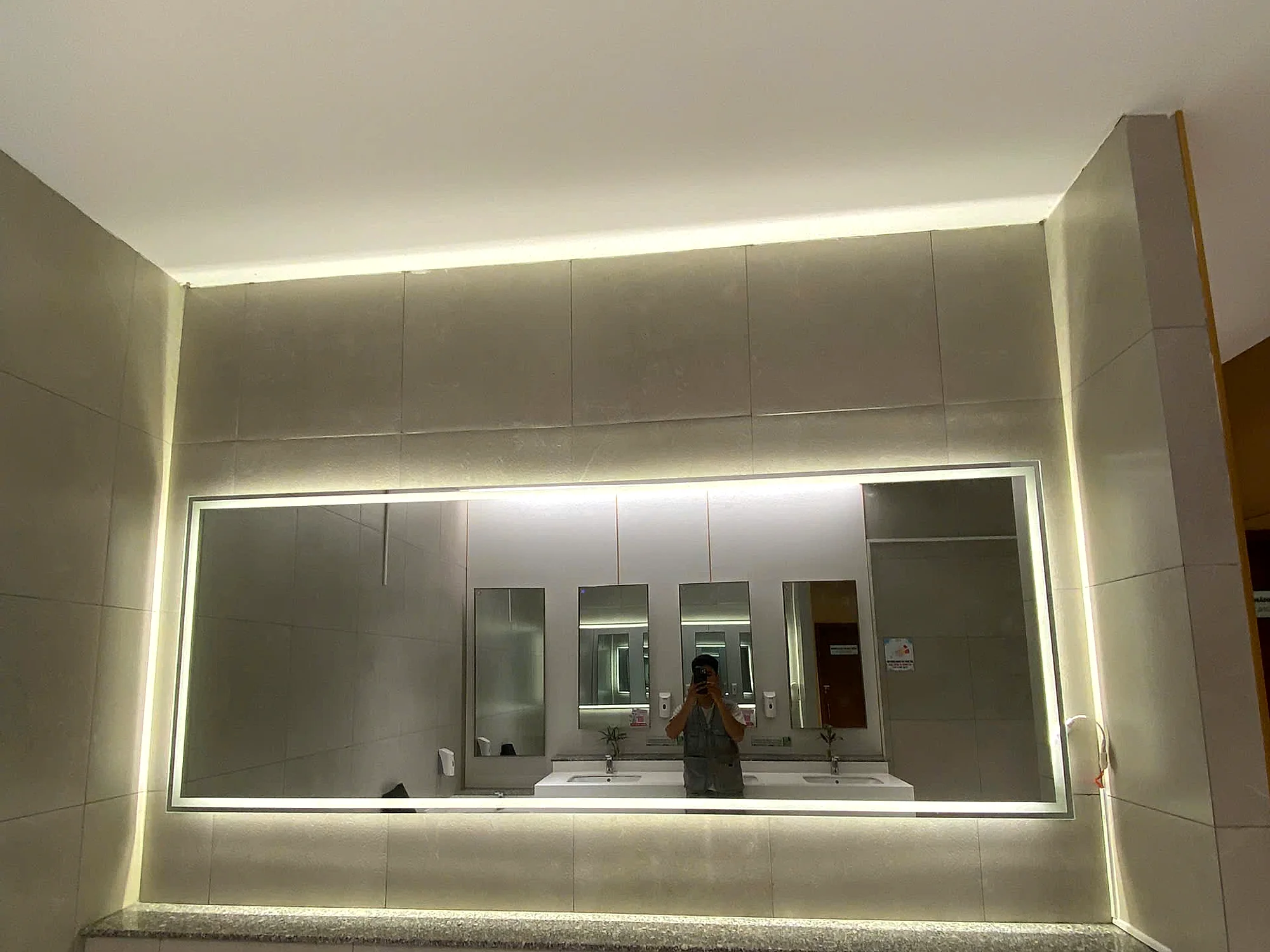 Gương soi khổ lớn tại khu vệ sinh công cộng PARC MALL, tường ốp đá màu be, viền LED ấm, ví dụ loại gương phù hợp cho khu vực ẩm và lưu lượng cao trong trung tâm thương mại