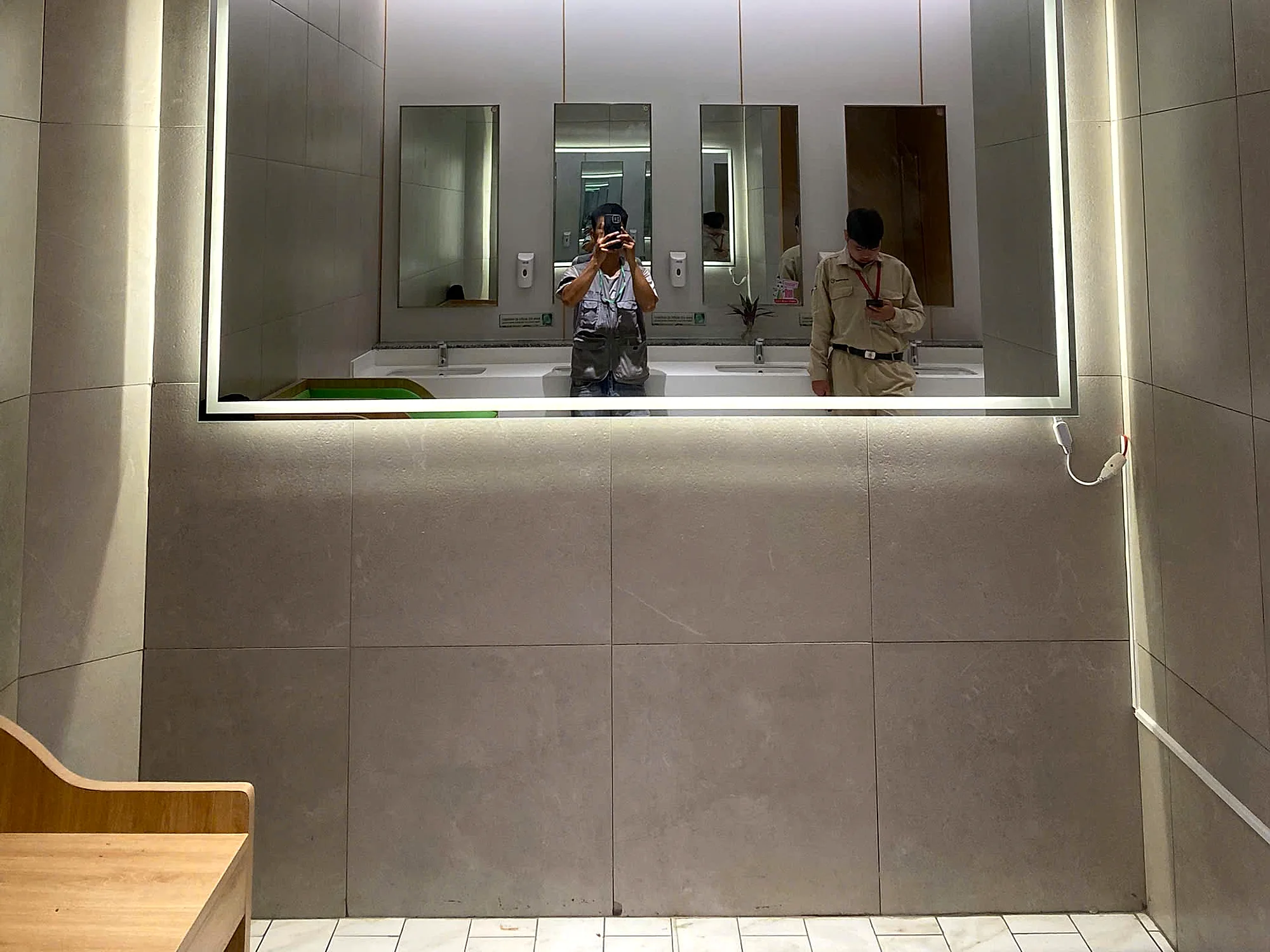 Khu vệ sinh PARC MALL với gương LED khổ lớn và ghế gỗ, tường ốp đá xám đã thi công hoàn thiện, viền đèn LED hắt sáng
