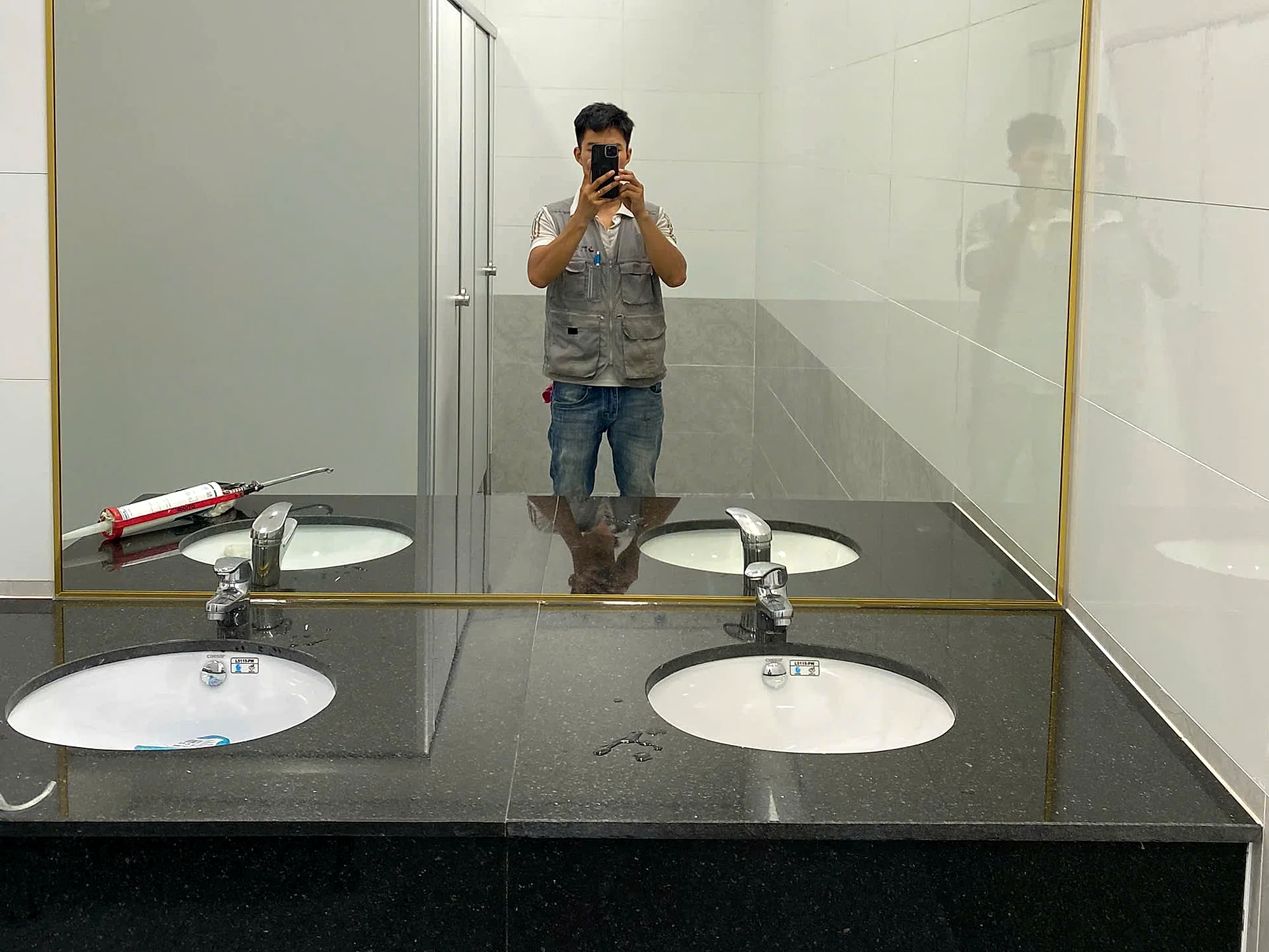 Công trình gương WC thực tế tại Hố Nai Đồng Nai, gương soi khung nhôm mạ vàng lắp trên bàn đá granite đen với hai lavabo sứ trắng, tường ốp gạch trắng viền hoa