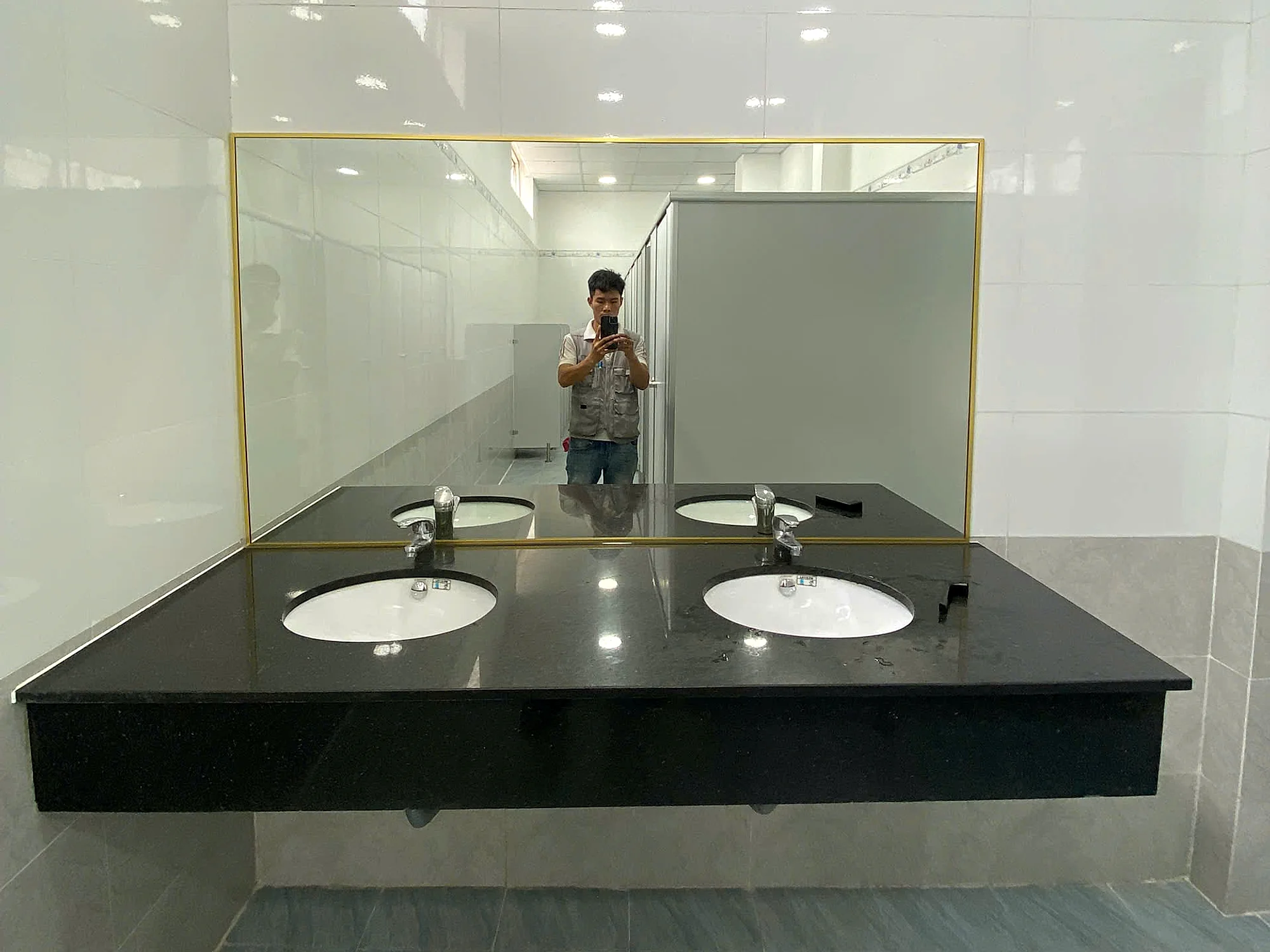 Mẫu gương nhà vệ sinh đã thi công tại Hố Nai, gương chữ nhật khung vàng trên bàn đá đen, nền gạch xám, ảnh công trình thực tế đã bàn giao