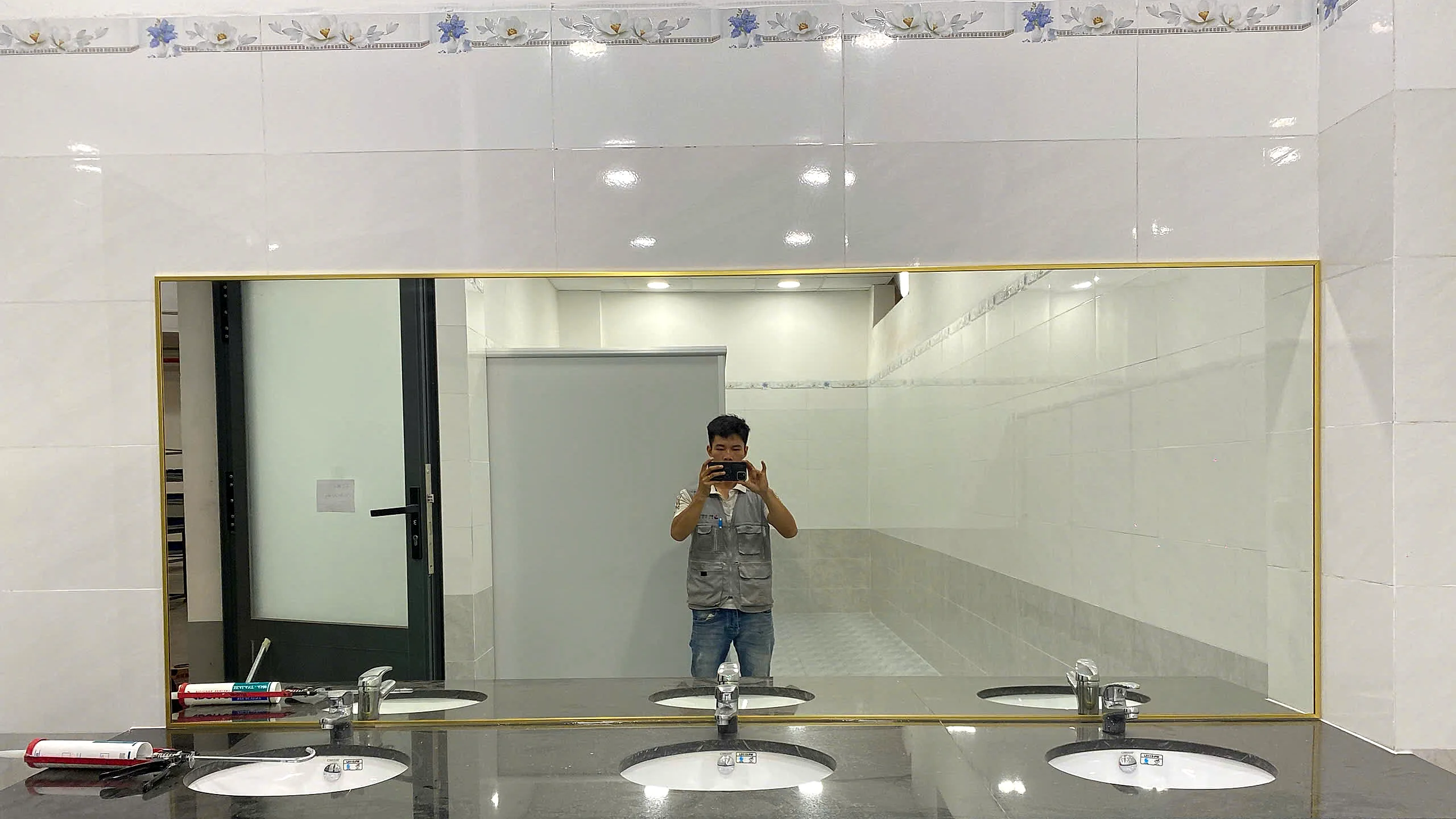 Công trình gương WC đã thi công hoàn thiện tại phường Hố Nai Đồng Nai, gương soi khung nhôm mạ vàng khổ lớn lắp trên bàn đá granite đen với dãy lavabo sứ trắng, tường ốp gạch sáng, ảnh thực tế tại nhà vệ sinh