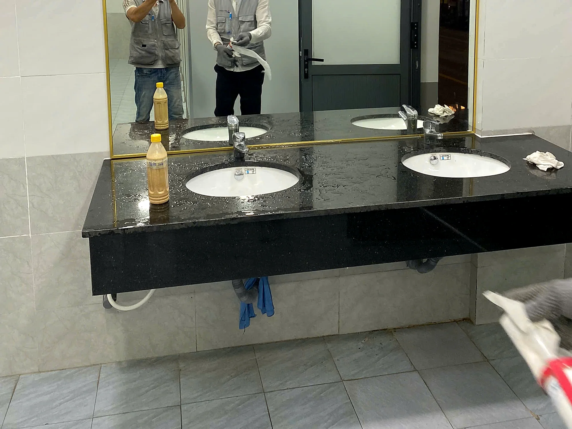 Thợ đang thi công lắp đặt gương WC tận nơi tại Hố Nai, đeo găng tay lau hoàn thiện tấm gương khung nhôm mạ vàng vừa lắp trên bàn đá granite đen, tuýp keo silicon trung tính đặt trên mặt bàn, không gian nhà vệ sinh thực tế đang hoàn thiện