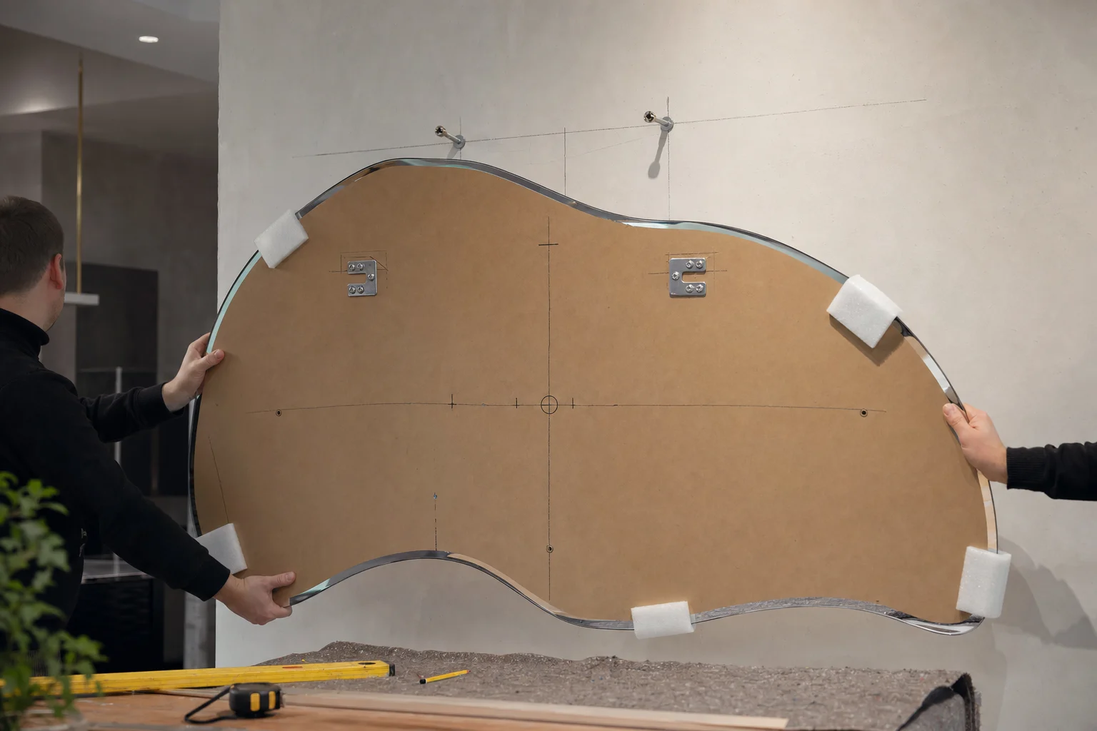 A realistic installation preparation scene for an irregular freeform mirror being mounted level on a wall in a professional glass workshop or on-site interior setting. Focus on the challenge of balancing an asymmetrical mirror: the visual center differs from the hanging center. Show the back of the mirror with carefully marked mounting points, center-of-gravity reference marks, top orientation marks, hanging brackets, and protective corner pads before delivery. Include polished mirror glass, irregular contour, wall anchors, measuring tools, and a partially finished modern interior wall. Soft technical lighting, close three-quarter camera angle, highly detailed craftsmanship, photorealistic, no text, no logo, no identifiable faces.