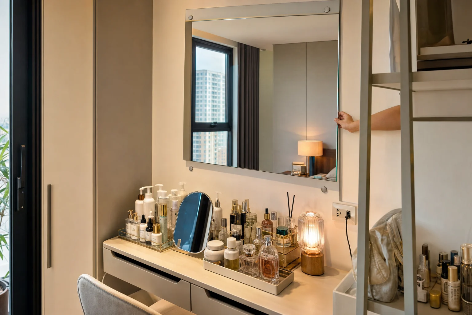 A realistic photo of a compact vanity with a shallow tabletop crowded by skincare bottles, perfume, trays, and a small lamp, plus an obstructing side shelf and wall outlet, illustrating why a full wall-mounted mirror is the better solution. Show the old small tabletop mirror pushed awkwardly among objects, while a larger rectangular wall mirror is being test-fitted above the vanity area. Emphasize mirror craftsmanship: clear silver mirror glass, slim aluminum frame or frameless polished edge option, secure wall mounting points, neat spacing above the table. Tight urban apartment setting in TP.HCM style, warm daylight mixed with soft interior light, camera angle from front three-quarter view to reveal the lack of usable depth on the tabletop. No readable text, no logo, no brand, no identifiable person, strictly mirror/glass-focused renovation scene.