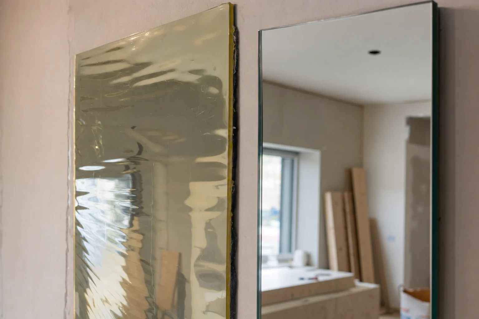 A close-up realistic comparison of mirror materials during installation, showing a flexible plastic adhesive mirror sheet on a wall with visible waviness, distorted reflection, slight yellow tint, and uneven surface, next to a true silver-backed glass mirror panel with sharp reflection and neutral color. Focus on the material difference causing image warping, ripples, and yellowing, not just bad mounting. Include mirror thickness contrast, exposed edge detail, backing material, construction setting, soft indoor lighting, oblique camera angle to highlight distortion, no text, no logo, no brand, no recognizable people.