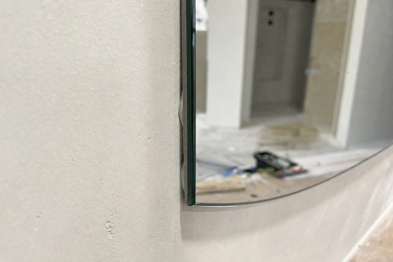 Photorealistic close-up of a large frameless mirror being installed on an uneven plaster wall, one section of the mirror edge sitting tight while another section reveals a visible tapered gap between mirror and wall, construction adhesive dabs hidden behind the glass, subtle wall waviness and imperfect substrate clearly visible, natural workshop-to-bathroom transition setting, side lighting to accentuate the gap and shadow line, macro architectural detail shot, realistic polished mirror edge, no text, no logo, no brand, no identifiable people