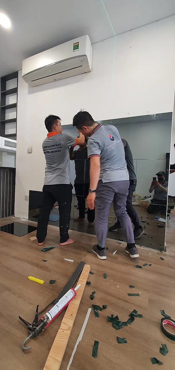 Xưởng Gương TP.HCM: Nhận Đơn Công Trình Và Số Lượng Lớn 2 Hai thợ nâng tấm gương lớn áp lên tường trong căn hộ đang hoàn thiện